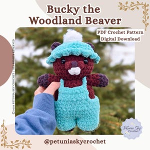 Op de afbeelding: Een handgemaakte gehaakte bever genaamd "Bucky" met een teal hoed en overall. Het pluche speelgoed is voornamelijk bordeauxrood met teal accenten en een witte pompon op de hoed. De afbeelding bevat tekst: "Bucky the Woodland Beaver".