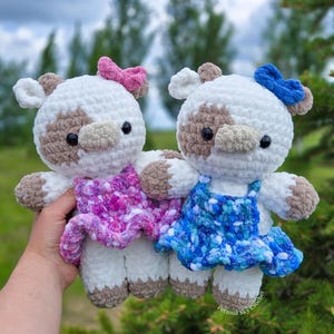 May include: Two handmade crochet cow plushies with white and tan bodies, black eyes, and tan snouts. One cow wears a pink dress and bow, while the other wears a blue dress and bow. The plushies feature soft, textured yarn and are held together.