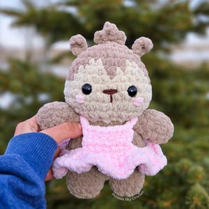May include: A handmade crochet squirrel doll in shades of brown, beige, and pink. The doll has a pink dress and black eyes. The text "Petunia Sky Crochet" is visible on the bottom right.