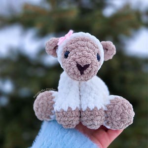 May include: A handmade crochet lamb toy. It has a white body, brown head, ears, and feet, and a pink bow. The lamb is sitting, with its legs and feet visible. The background is blurred, with green and white tones.