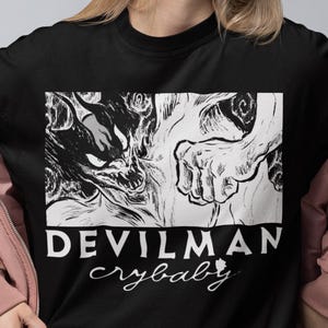 May include: Black t-shirt featuring a graphic from the anime series Devilman Crybaby. The design is in a white rectangular box and includes a detailed illustration of a demon and the text "DEVILMAN crybaby".