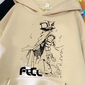 Puede incluir: Sudadera con capucha beige con bolsillo de canguro y capucha con cordón. La parte delantera presenta un gráfico en blanco y negro de una figura con la palabra "FECL" y "FOOLF COOLF" impresas debajo. La sudadera está hecha de un material suave.