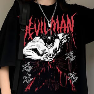 May include: Black t-shirt with the word "DEVILMAN" in red gothic lettering above a black and white illustration of a muscular figure. The shirt has short sleeves and a relaxed fit.