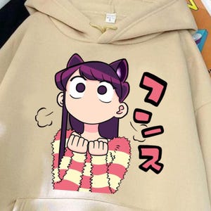 May include: A beige hooded sweatshirt featuring a graphic print of an anime character with dark purple hair and cat ears. The character wears a pink and yellow striped sweater. To the right of the character are large pink Japanese characters.