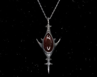 May include: A silver pendant necklace with a dark red gemstone in the center. The pendant is shaped like a stylized, pointed, abstract design.