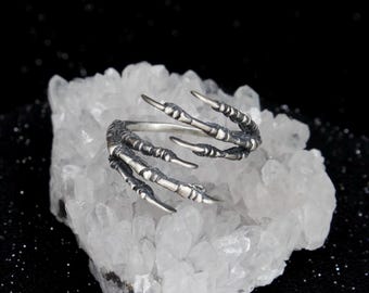 May include: A silver-toned ring shaped like a bird's claw with textured, segmented details. The metal band curves to form the talon, featuring darkened recessed areas that highlight the sharp, pointed tips of the claw design.