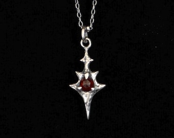 May include: A silver-toned metal pendant necklace featuring a sharp, multi-pointed star or cross-like shape with a textured, organic finish. A single, round, faceted deep red gemstone is set securely in the center of the metallic pendant.