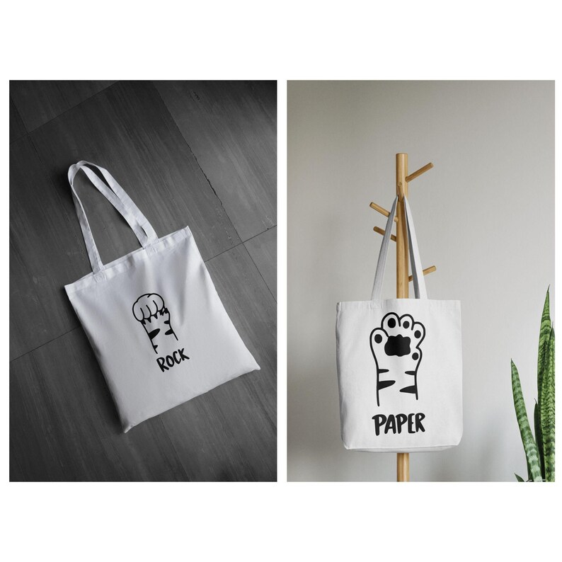 May include: White tote bags with black paw print designs and text. One bag says "ROCK" and the other says "PAPER". The bags have long handles and are made of a sturdy fabric.