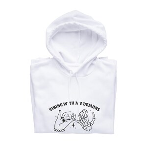 May include: White hooded sweatshirt with a black graphic print of two hands holding pinky fingers. The text "VIBING WITH MY DEMONS" is printed in black above the hands.