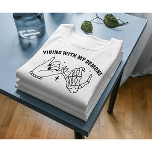 May include: White t-shirt with a black graphic of two hands holding pinky fingers. The text "VIBING WITH MY DEMONS" is printed on the shirt.