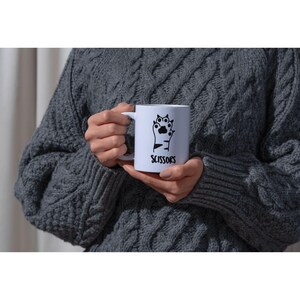 May include: White ceramic mug with a black graphic of a cat paw holding scissors. The word "SCISSORS" is printed below the graphic in black. The mug is held by a person wearing a gray cable-knit sweater.