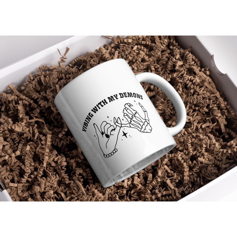 May include: White ceramic mug with a black design of two hands holding pinky fingers. The text on the mug reads "Vibing with my demons".
