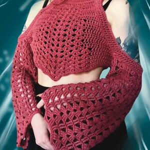 May include: A maroon crochet top with openwork detailing. The top features a high neckline, open shoulders, and long, flared sleeves. The handmade garment is a unique piece of apparel.