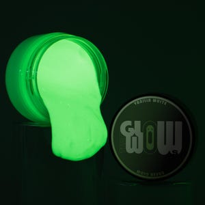 May include: A jar of green glow-in-the-dark slime with the lid off, spilling out onto a surface. The lid of the jar is next to the jar, with the words "The Glow" and "Vanilla White" printed on it.