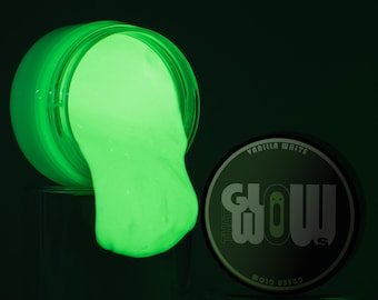 Könnte beinhalten: Ein Glas grünen, im Dunkeln leuchtenden Schleims, dessen Deckel geöffnet ist und auf eine Oberfläche verschüttet wird. Der Deckel des Glases befindet sich neben dem Glas, mit den Worten "The Glow" und "Vanilla White" darauf.
