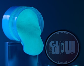 Könnte beinhalten: Ein Glas mit ozeanblauem, im Dunkeln leuchtendem Schleim, der auf eine blaue Oberfläche gegossen wird. Das Glas hat ein schwarzes Etikett mit der Aufschrift "That Glow" und "Sky Glow" sowie einer Geistergrafik.