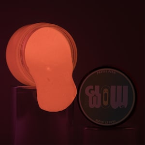Può includere: Un barattolo di slime rosa pastello che brilla al buio con la scritta "GLOW" e "MOJO TREASURES" sul coperchio.
