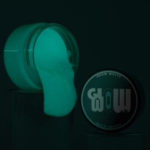 Peut inclure: Un pot de slime qui brille dans le noir de couleur vert turquoise. Le slime déborde du pot. Le pot a un couvercle noir avec les mots "The Glow" et "Snow White" imprimés dessus.