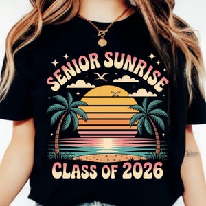 K&ouml;nnte beinhalten: Schwarzes T-Shirt mit einem farbenfrohen Grafikdesign. Das Design zeigt die Worte "SENIOR SUNRISE" in einer Retro-Schriftart &uuml;ber einer Sonnenuntergangsszene mit Palmen, Wolken und dem Text "CLASS OF 2026".