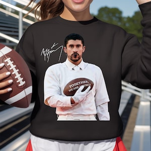 May include: Black sweatshirt featuring a portrait of a person holding a football. The football has the text "TOGETHER, WE ARE AMERICA". The person's signature is above the portrait. The sweatshirt is a crew neck style.