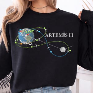 May include: Black sweatshirt featuring a graphic of Earth, the Moon, and a flight path with the text "ARTEMIS II" in white. The design includes a detailed illustration of the Earth with numbered points and a trajectory path.