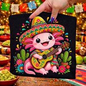 Pode incluir: Saco tote preto com um axolotl de desenho animado usando um chapéu e tocando guitarra. O axolotl é rosa e usa um poncho colorido. Cactos e flores cercam o axolotl. O fundo está desfocado com decorações festivas.