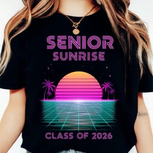 K&ouml;nnte beinhalten: Schwarzes T-Shirt mit Retro-Design. Der pinkfarbene Text "SENIOR SUNRISE" steht &uuml;ber einer Sonnenuntergangsgrafik. Darunter steht der Text "CLASS OF 2026". Das Design umfasst Palmen und ein Rastermuster, das an die 1980er Jahre erinnert.