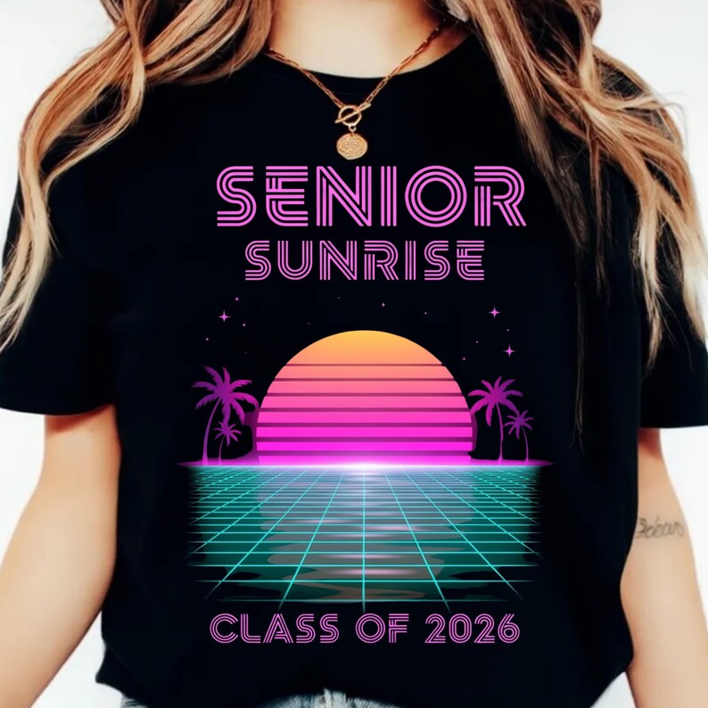 K&ouml;nnte beinhalten: Schwarzes T-Shirt mit Retro-Design. Der pinkfarbene Text "SENIOR SUNRISE" steht &uuml;ber einer Sonnenuntergangsgrafik. Darunter steht der Text "CLASS OF 2026". Das Design umfasst Palmen und ein Rastermuster, das an die 1980er Jahre erinnert.