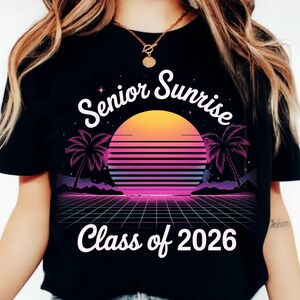 K&ouml;nnte beinhalten: Schwarzes T-Shirt mit Retro-Design, das einen Sonnenuntergang, Palmen und ein Gittermuster zeigt. Der Text "Senior Sunrise" w&ouml;lbt sich &uuml;ber der Sonne, darunter "Class of 2026". Das Design ist in Orange-, Rosa-, Lila- und Blaut&ouml;nen gehalten.