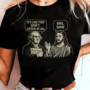 May include: Black t-shirt featuring a graphic of George Washington and Jesus Christ in a humorous conversation. Washington holds a document that reads "We the People." Speech bubbles above them say "It's like they didn't listen at all" and "Bro, Same."
