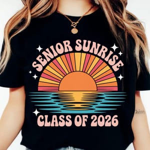 K&ouml;nnte beinhalten: Schwarzes T-Shirt mit einem Retro-Sonnenuntergangsdesign. Die Worte "SENIOR SUNRISE" w&ouml;lben sich &uuml;ber einer Sonnengrafik, darunter "CLASS OF 2026". Der Sonnenuntergang zeigt orange, rosa und gelbe Strahlen &uuml;ber blauem Wasser. Wei&szlig;e Sternschnuppen erg&auml;nzen das Design.