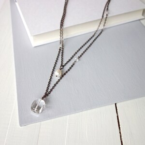 Puede incluir: Un delicado collar de dos capas con un colgante de cuarzo transparente y un dije de perla. El collar está hecho de cadena de cobre y presenta pequeños detalles de cristal.