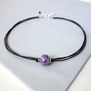 May include: A necklace featuring multiple thin black cords and a single central purple bead with a glossy finish. The bead is flanked by two small silver-toned metal rings. The necklace includes a silver-toned metal chain extender at the back.