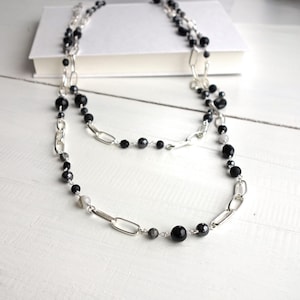 May include: A double-strand necklace with black beads and silver-coloured chain links. The necklace is presented on a white surface, with a white book in the background. The beads are of varying sizes and shapes, adding visual interest to the jewellery.