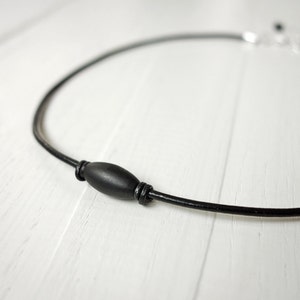 Black leather cord necklace with knots of leather o an each side of a black metal tube bead which is thicker in the middle.