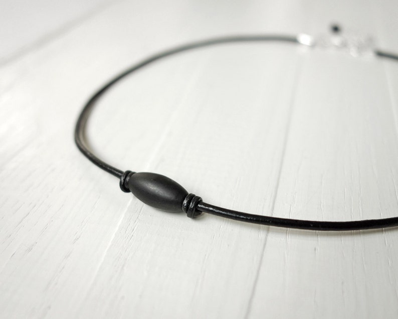 Black leather cord necklace with knots of leather o an each side of a black metal tube bead which is thicker in the middle.