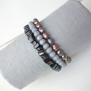 May include: Three bracelets stacked on a gray fabric surface. The top bracelet is made of gray and pink pearls. The middle bracelet is made of gray faceted beads. The bottom bracelet is made of black and silver metal beads.