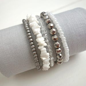 Könnte beinhalten: Ein Set aus sechs Armbändern mit verschiedenen Perlenstilen. Die Armbänder bestehen aus weißen Perlen, silbernen Perlen und kleinen grauen Perlen. Die Armbänder sind übereinander gestapelt.