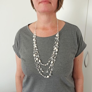 May include: A multi-strand necklace featuring clear crystals, white pearls, and silver-coloured metal rings. The necklace is worn over a grey t-shirt. The necklace has three strands of varying lengths.
