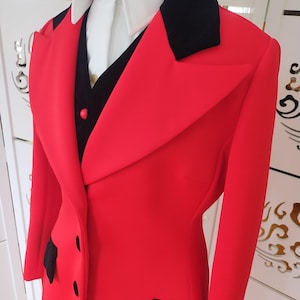 Può includere: Un blazer rosso acceso con dettagli neri, tra cui un colletto nero e dettagli delle tasche. Il blazer è abbinato a una camicia bianca con colletto e un gilet nero, creando un forte contrasto. La giacca ha tre bottoni neri.