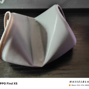 May include: A gray, folded, geometric object with a soft, textured appearance. The object has a unique, crumpled shape, with visible folds and creases. The lighting creates highlights and shadows, emphasizing the form and texture of the material.