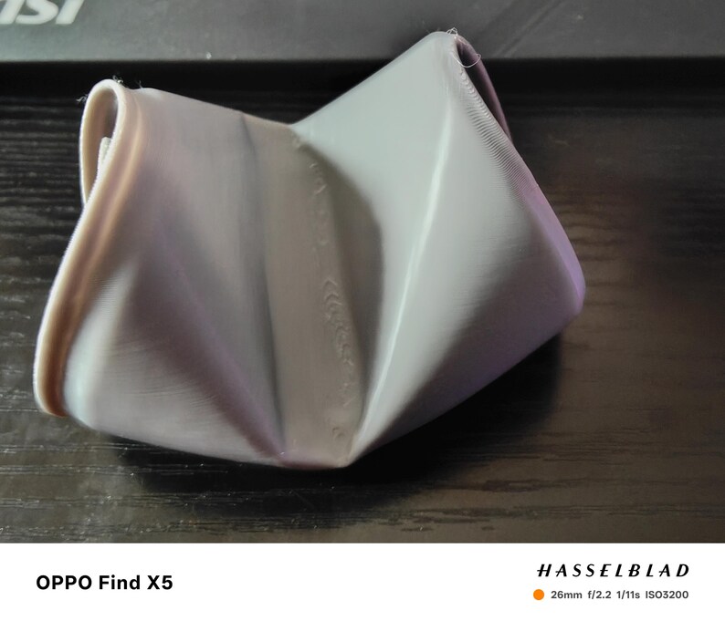 May include: A gray, folded, geometric object with a soft, textured appearance. The object has a unique, crumpled shape, with visible folds and creases. The lighting creates highlights and shadows, emphasizing the form and texture of the material.