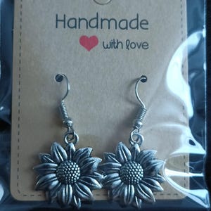 May include: A pair of silver-colored sunflower earrings with a detailed design. The earrings are displayed on a brown card that reads "Handmade with love". The earrings have a hook closure.