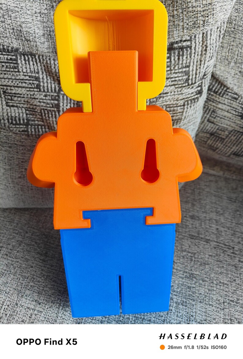 May include: A brightly colored, abstract 3D-printed figure in yellow, orange, and blue. The figure has a rectangular head, a torso with two keyhole-shaped cutouts, and a pair of arms. The lower portion is blue with a vertical slot. The text "OPPO Find X5" is at the bottom.