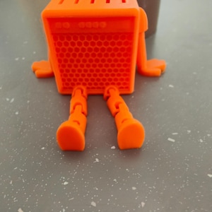 May include: An orange, stylized, 3D-printed object resembling an amplifier with legs and arms. The front of the amplifier has a honeycomb pattern. The top has several parallel slots. The object is on a gray surface.