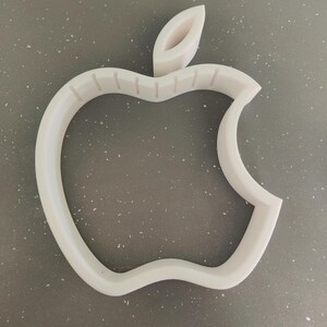 May include: A white, three-dimensional apple logo, with a missing bite, is centered in the image. The logo is made of a solid, opaque material and sits on a speckled gray surface. The stem is also white and three-dimensional.