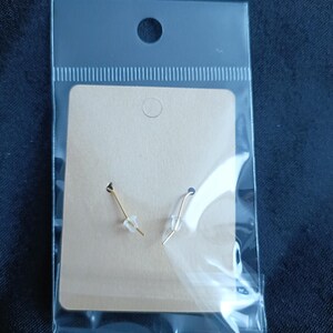May include: A pair of gold earrings with clear plastic stoppers, presented on a tan cardboard backing within a clear plastic package. The earrings have a simple, elegant design.