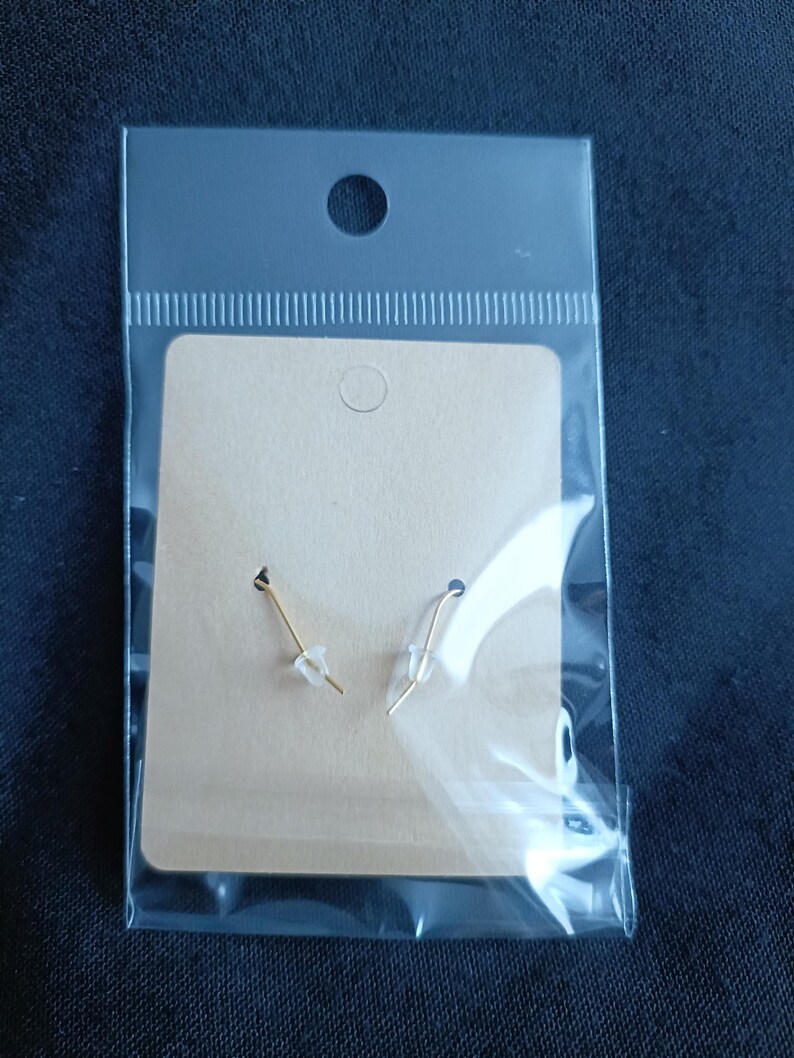 May include: A pair of gold earrings with clear plastic stoppers, presented on a tan cardboard backing within a clear plastic package. The earrings have a simple, elegant design.
