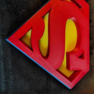 May include: A red, yellow, and blue emblem with a stylized "JP" design. The emblem features a red border with a blue outline, enclosing a yellow background with the letters "JP" in red. The design is set against a dark, textured background.