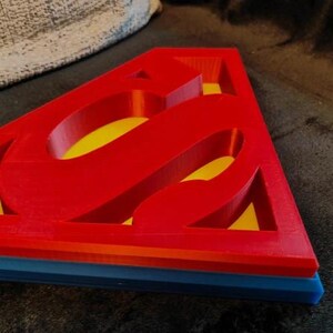May include: A red, yellow, and blue Superman logo. The iconic "S" emblem is prominently displayed in red, with a yellow interior. The base of the logo is blue, with orange and yellow layers. The item is likely a decorative piece or a toy.
